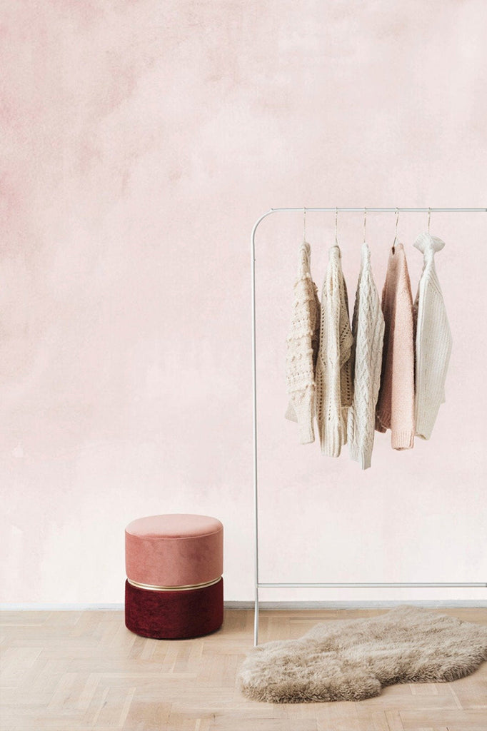 A cozy room featuring the Aista Ombre Watercolour Wallpaper with a  Blush Pink to cream gradient. A metal clothing rack holds knitwear, beside a plush round burgundy ottoman and a fluffy beige rug on light wooden flooring.