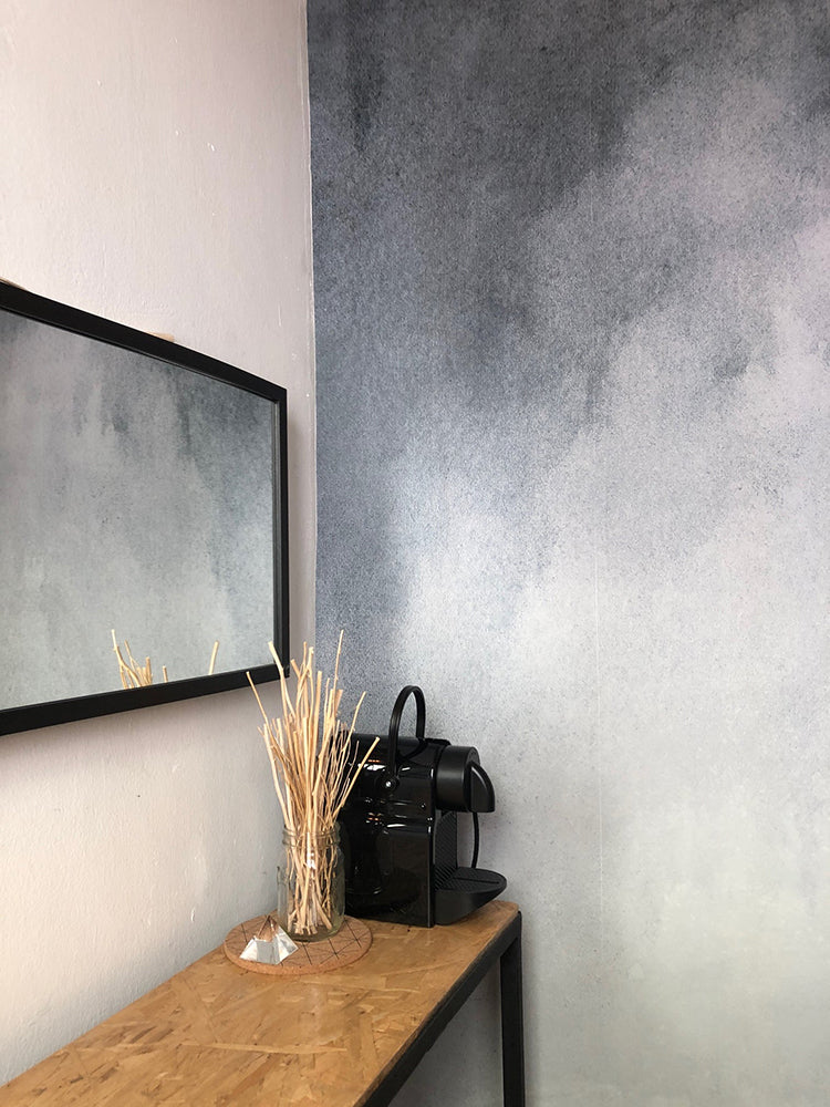 A serene room adorned with the Aista Ombre Watercolour Wallpaper in Grey. The gradient transitions from dark hues at the top to lighter shades near the bottom. A wooden console table stands against the wall, featuring a vase with dried plants and a black teapot set. A large mirror reflects the elegant ambiance.