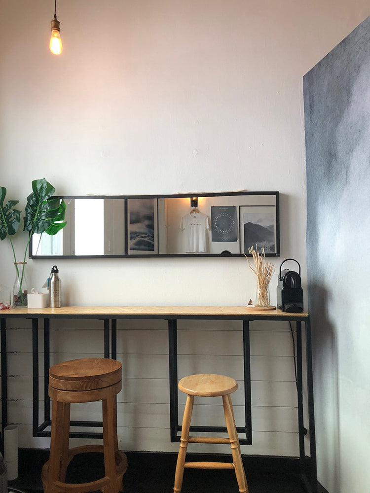 The modern room features a minimalist design with a white wall on the left. On the right, the Aista Ombre Watercolour Wallpaper blends from white to grey. A sleek black-framed mirror hangs above a simple console table adorned with decorative items. Two wooden stools flank the table, creating a balanced and inviting space.