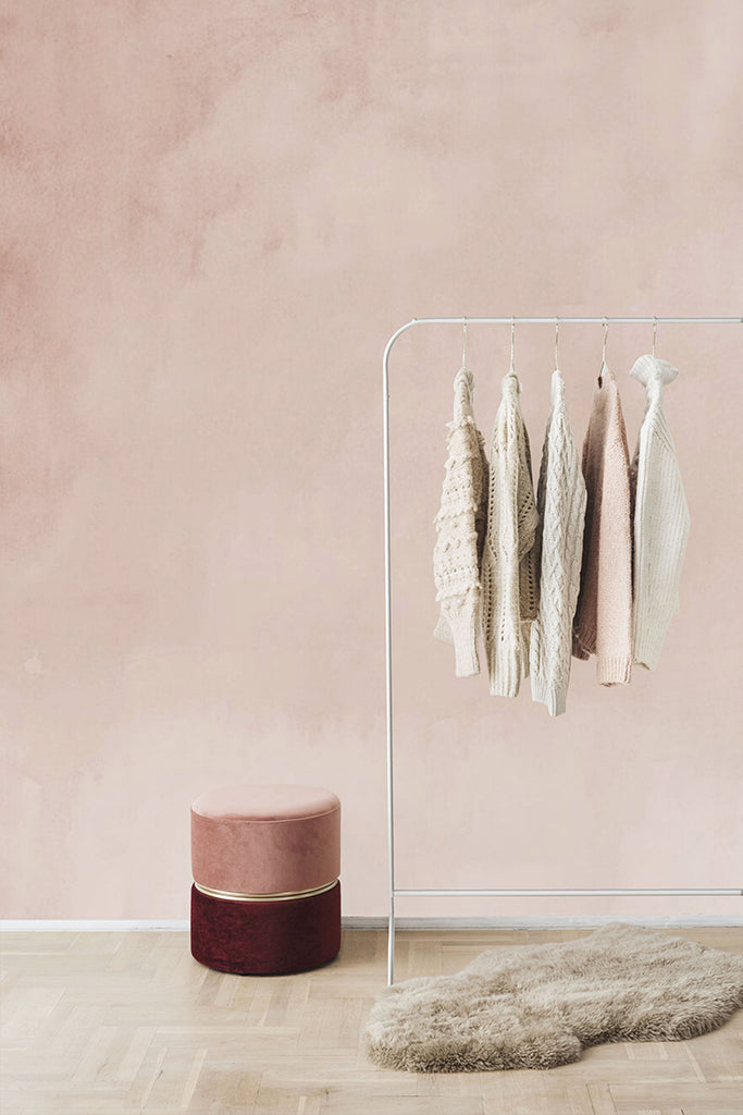 A cozy room featuring the Aista Ombre Watercolour Wallpaper with a  Nude to cream gradient. A metal clothing rack holds knitwear, beside a plush round burgundy ottoman and a fluffy beige rug on light wooden flooring.