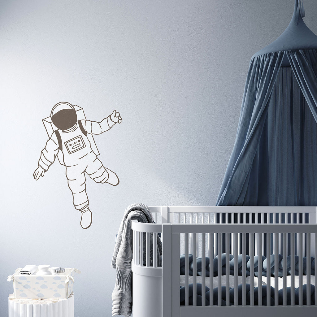 A serene nursery room with light gray walls adorned by a playful wall decal named Astronaut, Wall Decals. Beside it, a dark-rail crib sits under a draped canopy. A small, patterned hamper adds a touch of charm.