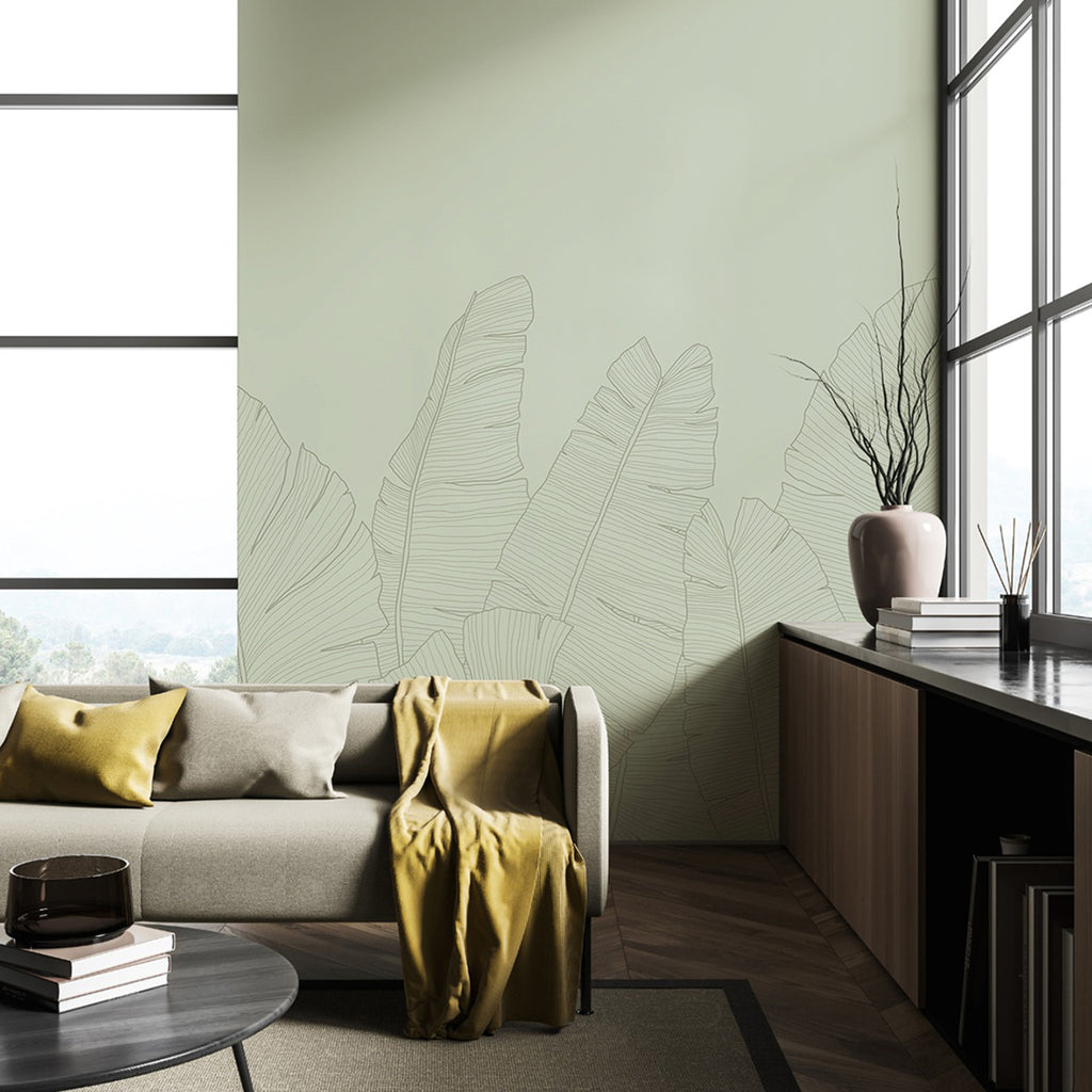 The room features large windows, a beige sofa adorned with yellow pillows, and a wooden floor. The wall showcases the Banana Foliage Tropical Mural Wallpaper, which depicts large, outlined banana leaves on a pale green background