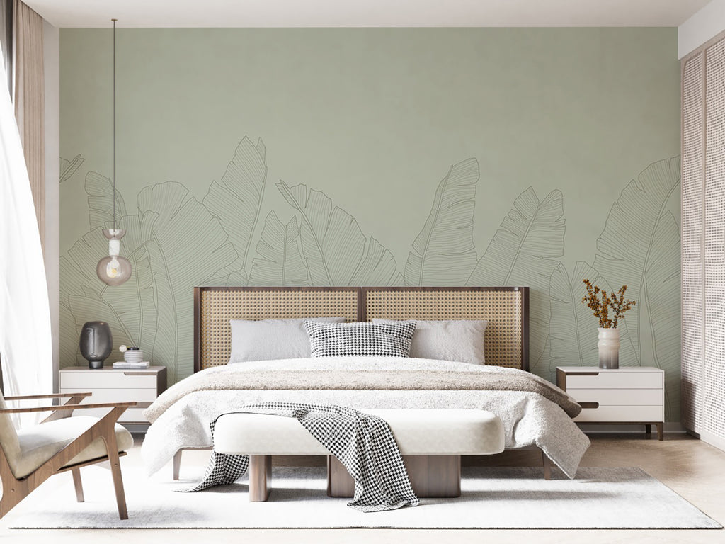 The room features large, Banana Foliage, Tropical Mural Wallpaper in Green. A modern bed with white bedding and a throw sits against the botanical backdrop. Wooden side tables and minimalist decor complete the tranquil space. A woven headboard adds texture to the room