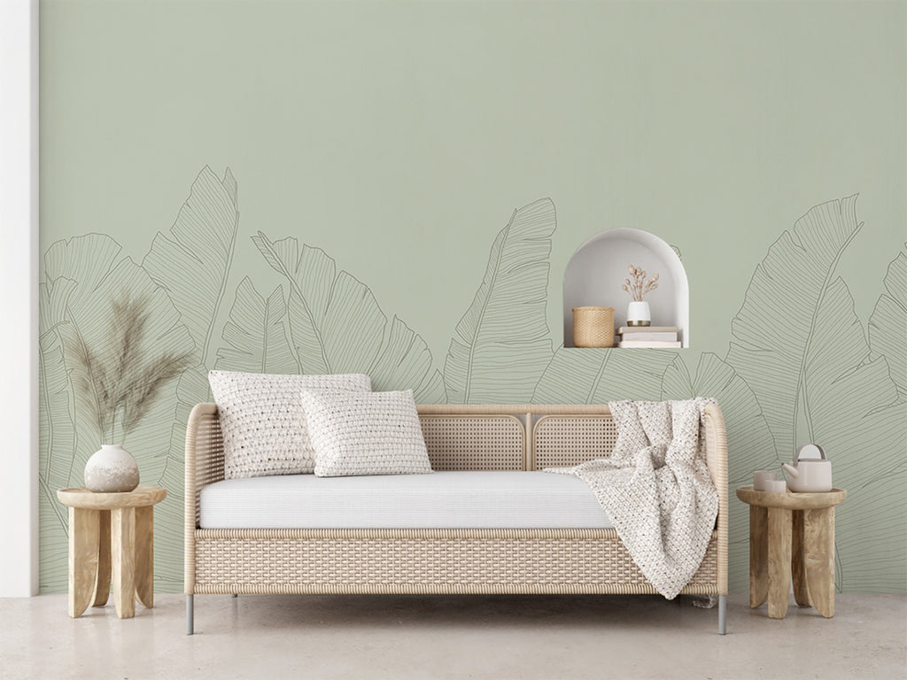 The room features a serene ambiance with a wall adorned by the Banana Foliage, Tropical Mural Wallpaper in Green. The wallpaper showcases large, detailed tropical leaves in a muted green hue. A modern beige sofa with white cushions and a cozy throw blanket sits in front of the wallpaper. Wooden side tables with decorative items and vase, creating an inviting and stylish space.