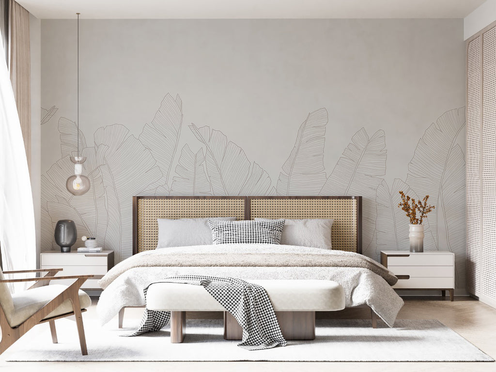 The room features large, Banana Foliage, Tropical Mural Wallpaper in Grey. A modern bed with white bedding and a  throw sits against the botanical backdrop. Wooden side tables and minimalist decor complete the tranquil space. A woven headboard adds texture to the room