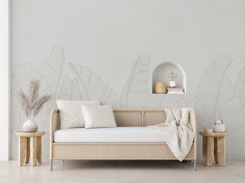 The room features a serene ambiance with a wall adorned by the Banana Foliage, Tropical Mural Wallpaper in Grey. The wallpaper showcases large, detailed tropical leaves in a muted Grey hue. A modern beige sofa with white cushions and a cozy throw blanket sits in front of the wallpaper. Wooden side tables with decorative items and a vase, creating an inviting and stylish space.