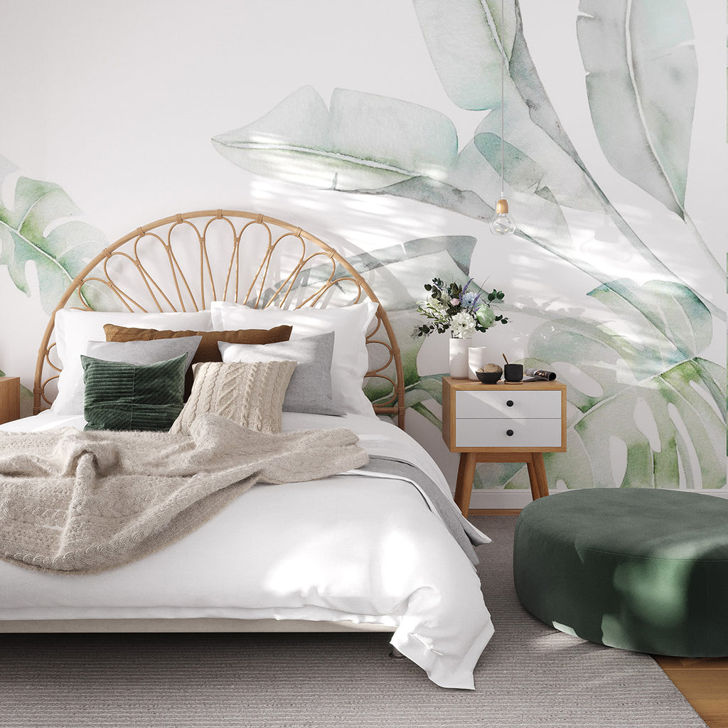 A serene bedroom adorned with the ‘Garden Bloom, Tropical Mural Wallpaper. The large green botanical prints create a calming atmosphere. The room includes a rattan headboard, white bedding, a desk with a plant, and toys scattered on the floor.