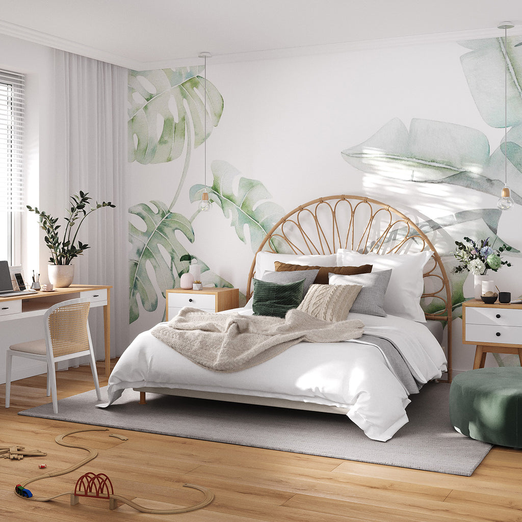 The ‘Garden Bloom, Tropical Mural Wallpaper adorns a serene bedroom. Its large green botanical patterns create a lively yet calming atmosphere. The room features a rattan headboard, white bedding, emerald green accents, and a modern bedside table with fresh flowers.