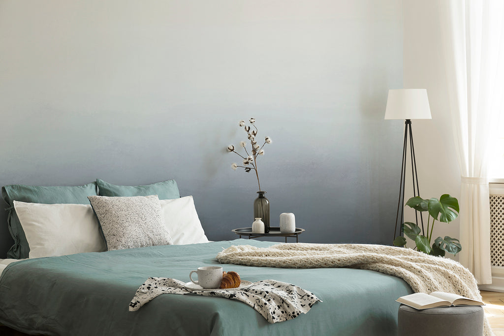 A serene bedroom with the Blake Horizon, Ombre Mural Wallpaper in Blue as the focal point. The room includes a cozy bed with teal bedding, a fluffy white throw, and a bedside table adorned with candles and a vase. A floor lamp and a potted plant add warmth and natural elements