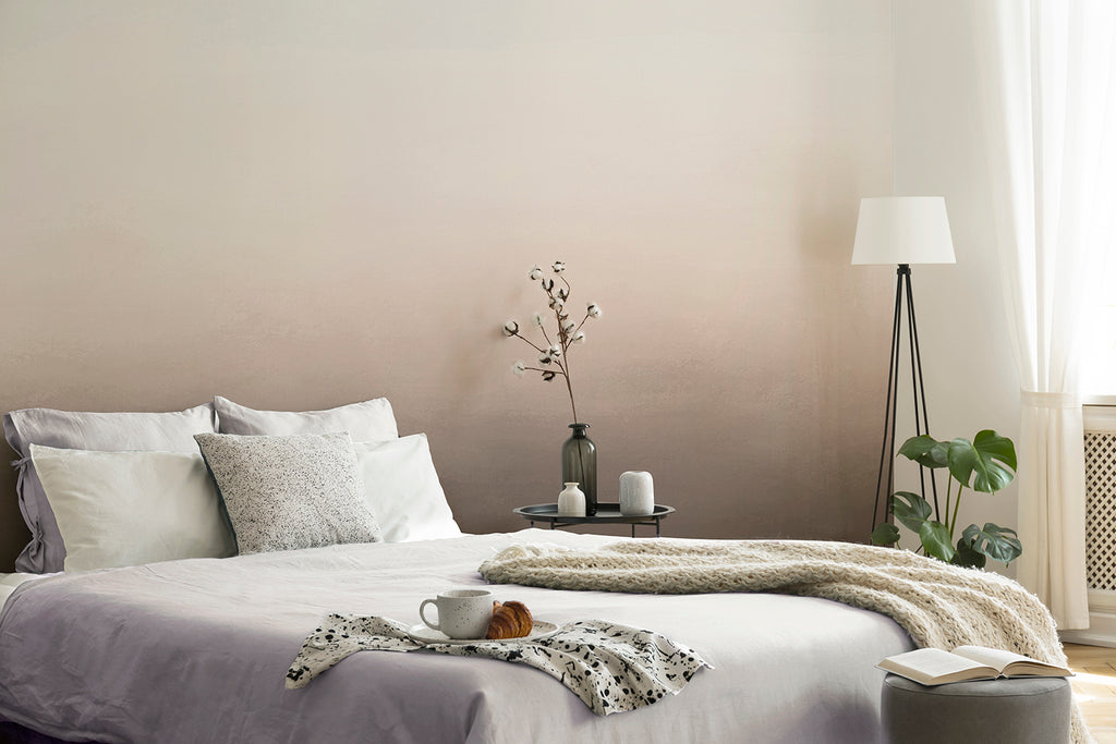 A serene bedroom with the Blake Horizon, Ombre Mural Wallpaper in Sand as the focal point. The room includes a cozy bed with teal bedding, a fluffy white throw, and a bedside table adorned with candles and a vase. A floor lamp and a potted plant add warmth and natural elements