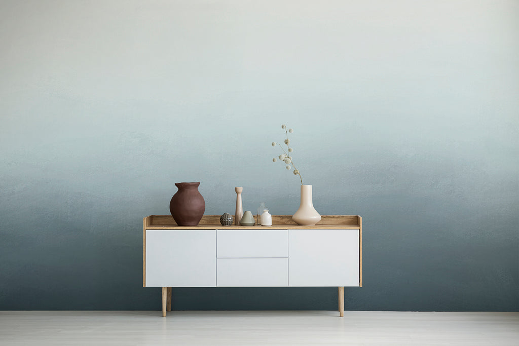 A minimalist room with a wooden console table against a wall featuring the Blake Horizon, Ombre Mural Wallpaper in Turquiose. Featuring various ceramic vases on a side board.