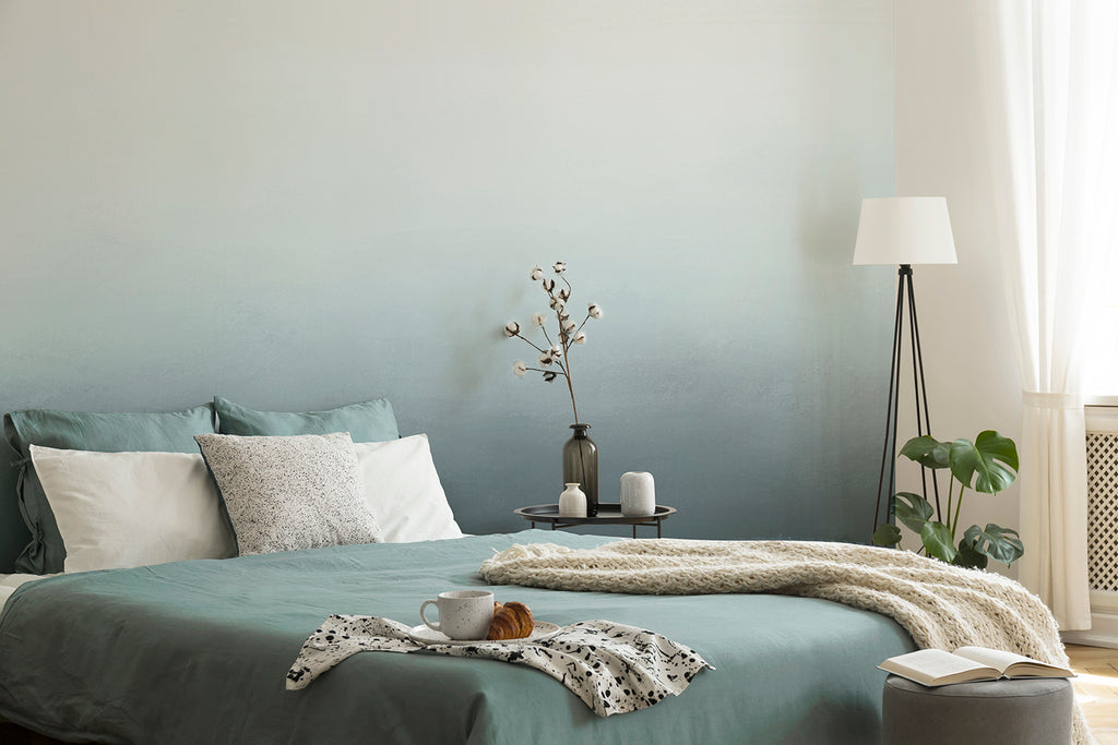 A serene bedroom with the Blake Horizon, Ombre Mural Wallpaper in Turquiose as the focal point. The room includes a cozy bed with teal bedding, a fluffy white throw, and a bedside table adorned with candles and a vase. A floor lamp and a potted plant add warmth and natural elements