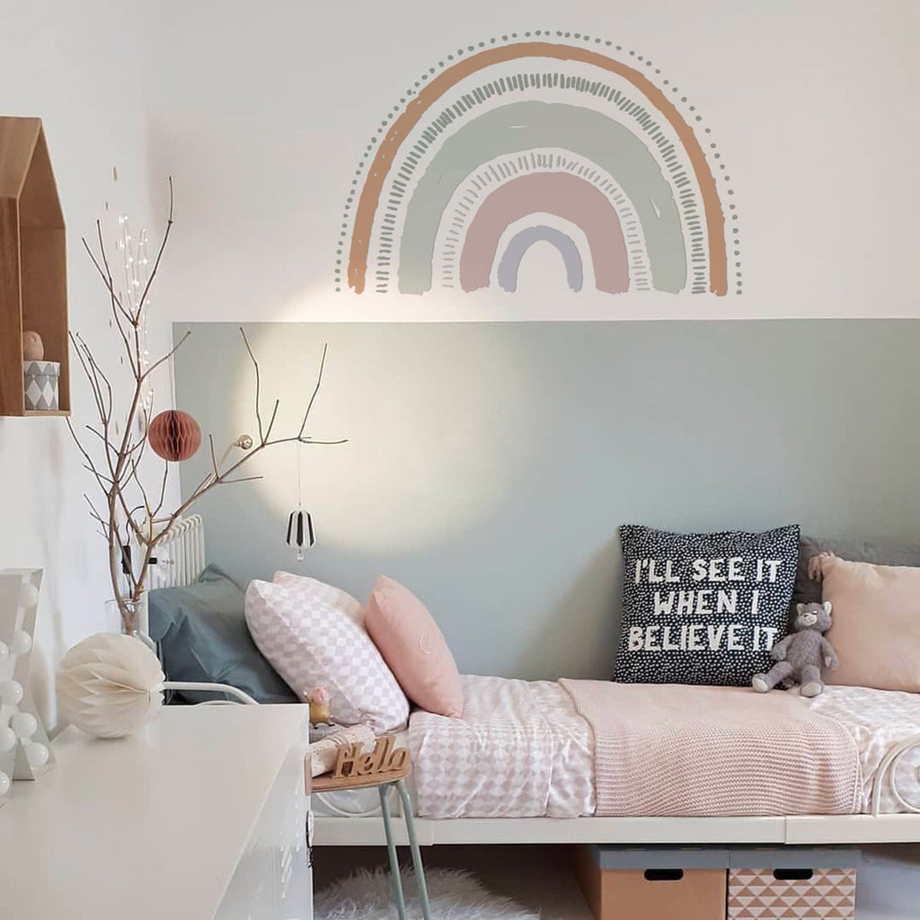 A cozy room featuring a pastel-colored Boho Large Rainbow, Wall Decals above a single bed with pink bedding. A white side table with a ‘hello’ sign, decorative vase, and hanging plant accentuate the serene ambiance.