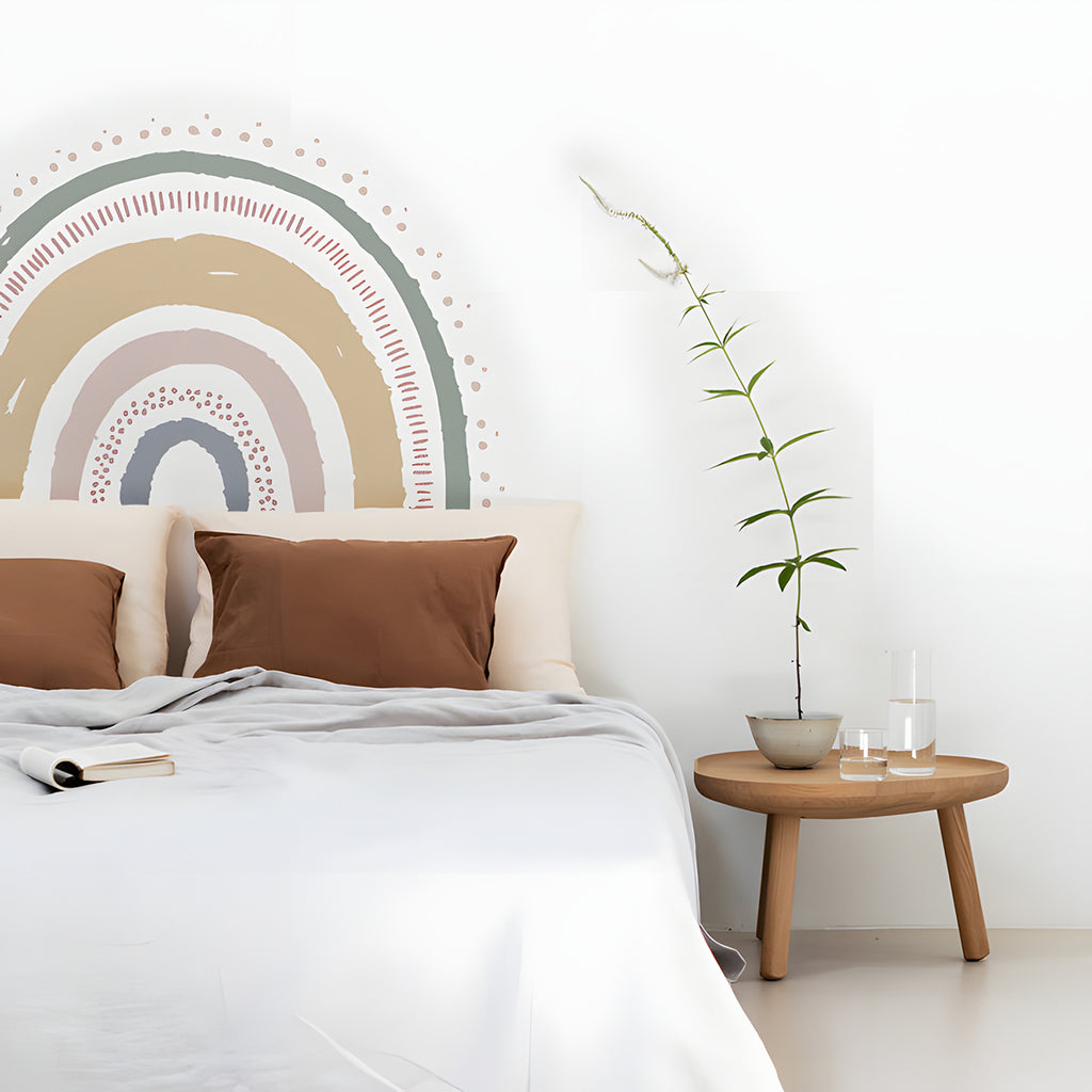 The room features a serene ambiance with minimalist design. The Boho XL Rainbow wall decals serve as an artistic focal point above a bed adorned with white linens and brown pillows. A wooden side table holds books and candles, while a tall green plant adds a touch of nature. 