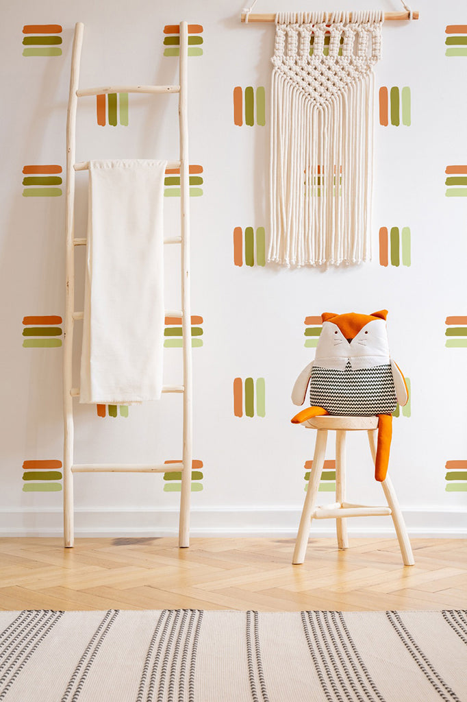 A cozy room with a white wall adorned by Bold Lines, Wall Decals in Light Green - Dark Green - Brown. A wooden ladder with a hanging cloth, an orange fox cushion on a chair, and woven wall art complete the warm decor.