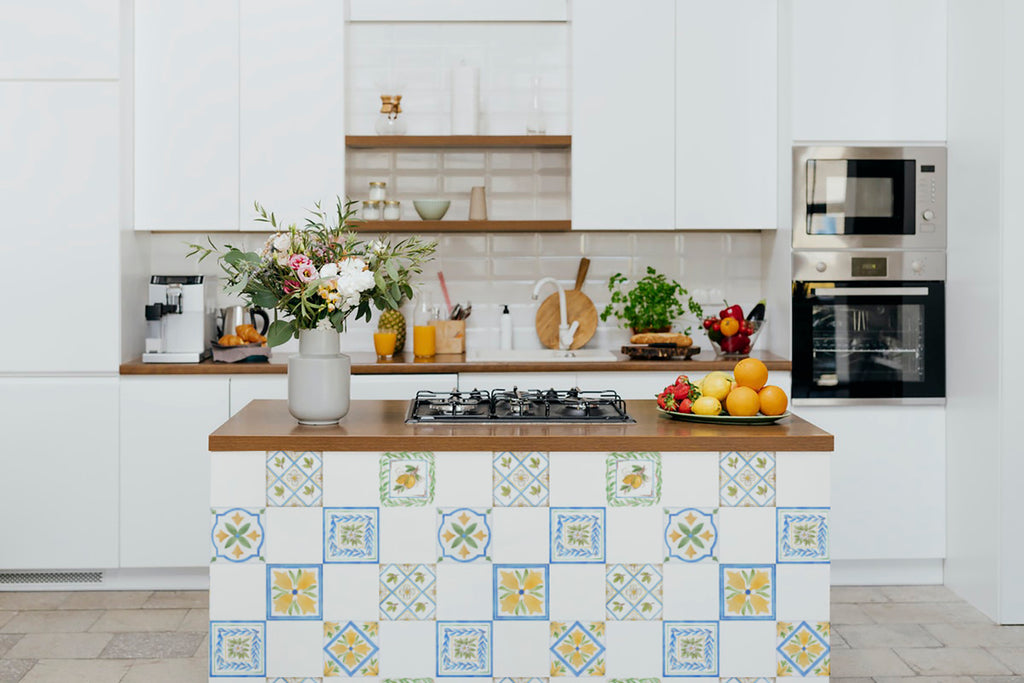 A stylish kitchen featuring Capri Tiles, Pattern Wallpaper. The vibrant mix of blue, yellow, and green designs on the tiles adds a pop of color. White countertops and neatly arranged utensils complete the modern look.