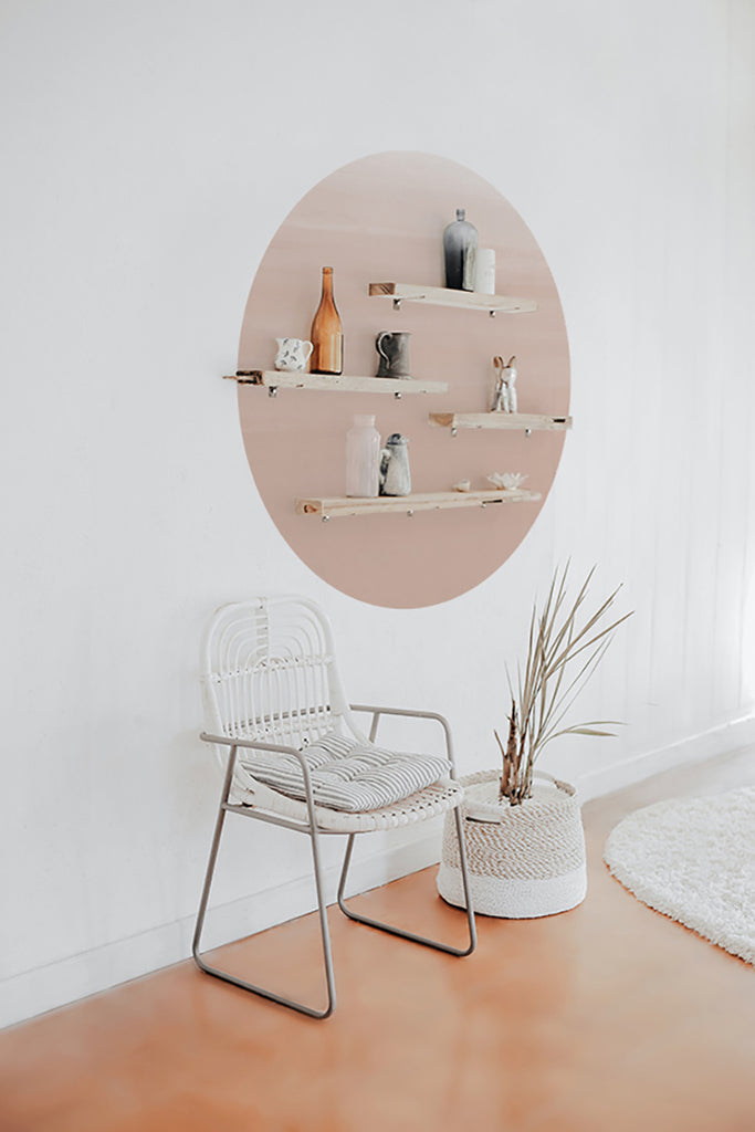 A modern room with a white chair and a Circle Ombre Gradient, Wall Decals in shades of blush pink. A white shelf with decorative items is mounted on the decal, complemented by a potted plant beside the chair.
