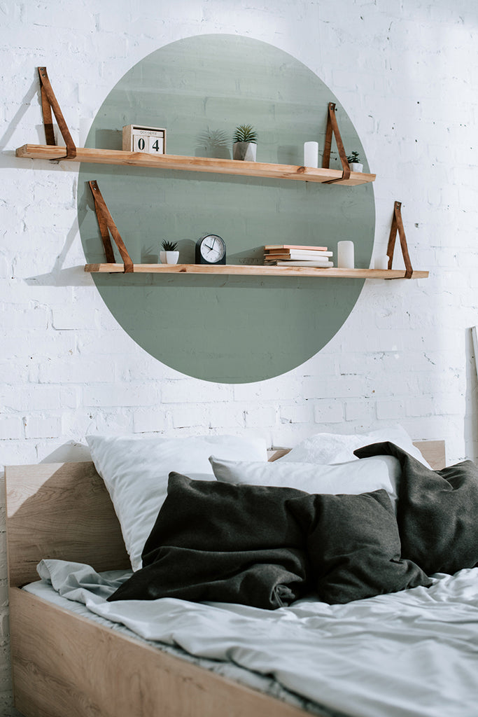A cozy bedroom with minimalist decor. The Circle Ombre Gradient, Wall Decals in Sage Green tones serves as the focal point. Above a bed with dark pillows, two wooden shelves hold plants, books, and decorative items against a white brick wall. The blend of modern design elements and comforting home decor creates a serene atmosphere.