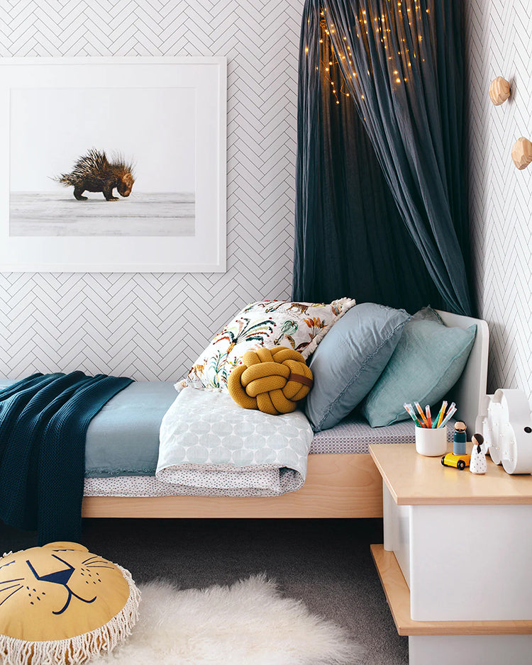 A cozy bedroom adorned with Classic Chevron Pattern Wallpaper in Black, featuring a single bed with blue bedding, a white-framed art piece, dark curtains with string lights, a white nightstand, and a yellow stool.