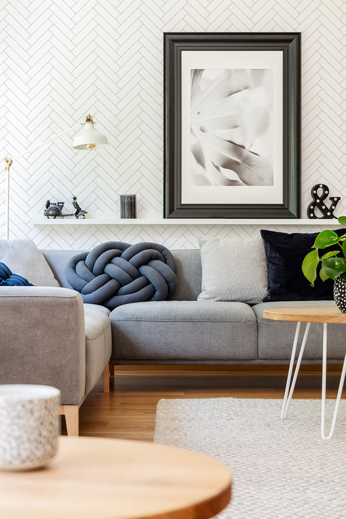 Modern living room featuring a Classic Chevron Patterned Wallpaper in Grey. The room is adorned with a grey sofa, textured cushions, gold-tone lighting fixtures, and decorative items on a side table, creating an elegant ambiance.