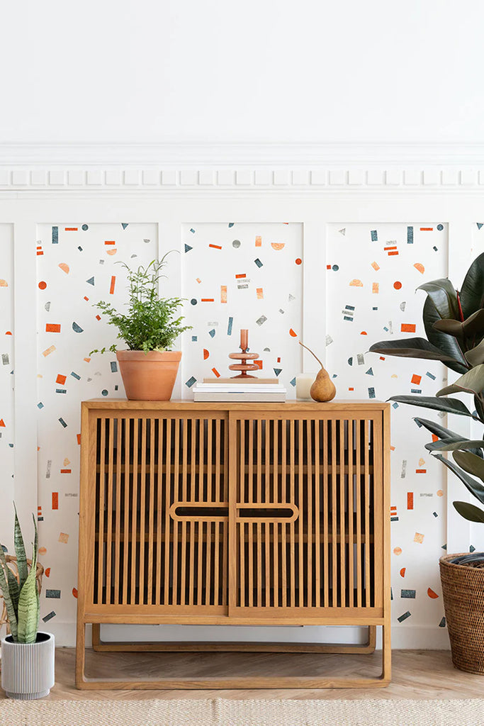 A contemporary room featuring a wooden cabinet against a vibrant wall. The wall is adorned with a Confetti Party, Pattern Wallpaper, filled with colorful geometric shapes in Crimson Dark Blue colourway. A potted plant adds a touch of nature atop the cabinet, and a large leafy plant to the right enhances the room’s serene.