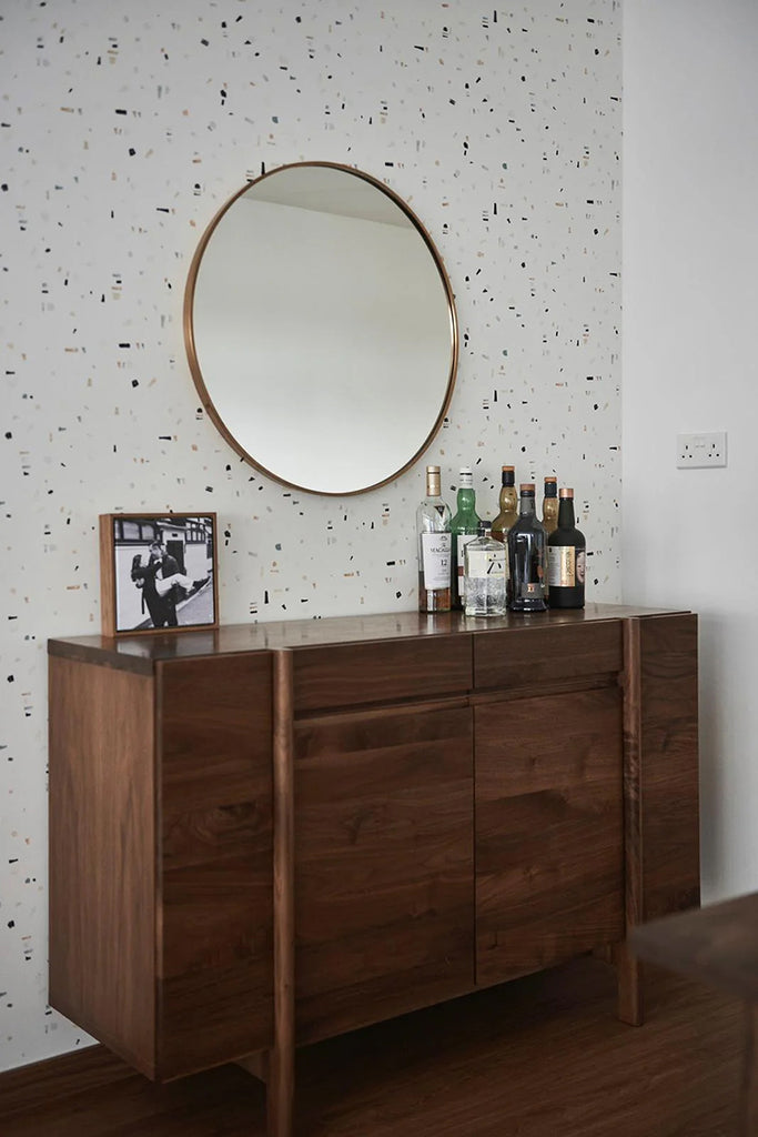 Modern room with a minimalist design, featuring a wooden sideboard adorned with various bottles and a framed photo. A large round mirror hangs above, all set against a playful Confetti Party, Patterned Wallpaper in Stratos Grey Blush Pink, adding a touch of whimsy to the space.