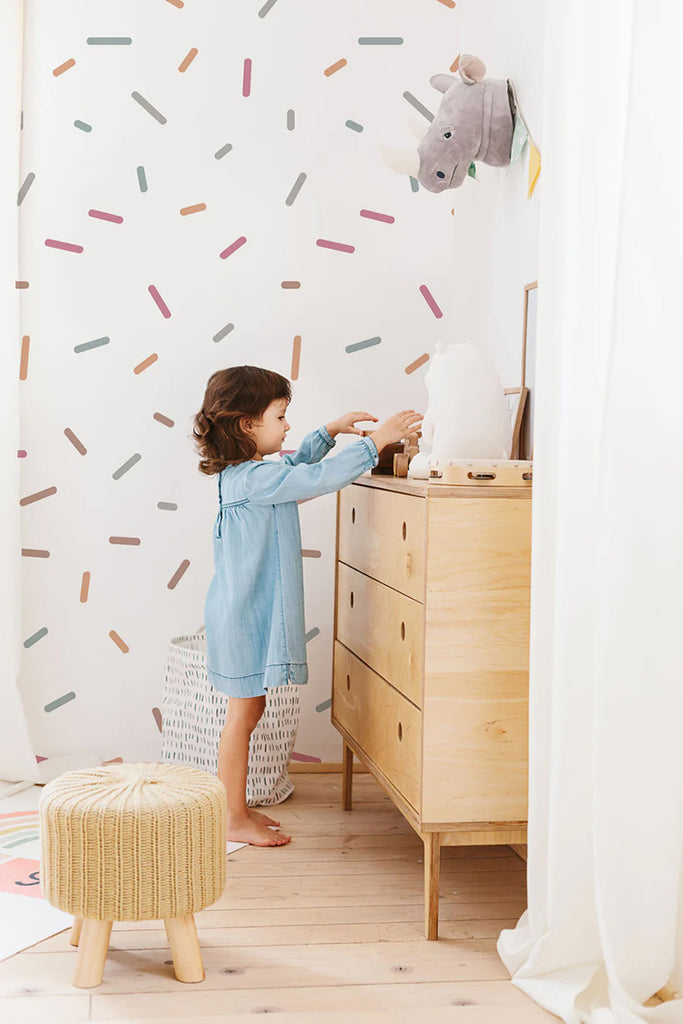 A lively child’s room with Confetti Shower, Pattern Wallpaper in Teal Honey, creating a vibrant backdrop. A child in a blue dress interacts with a wooden dresser, while a whimsical stuffed animal head adorns the wall. A small stool adds to the room’s playful charm.