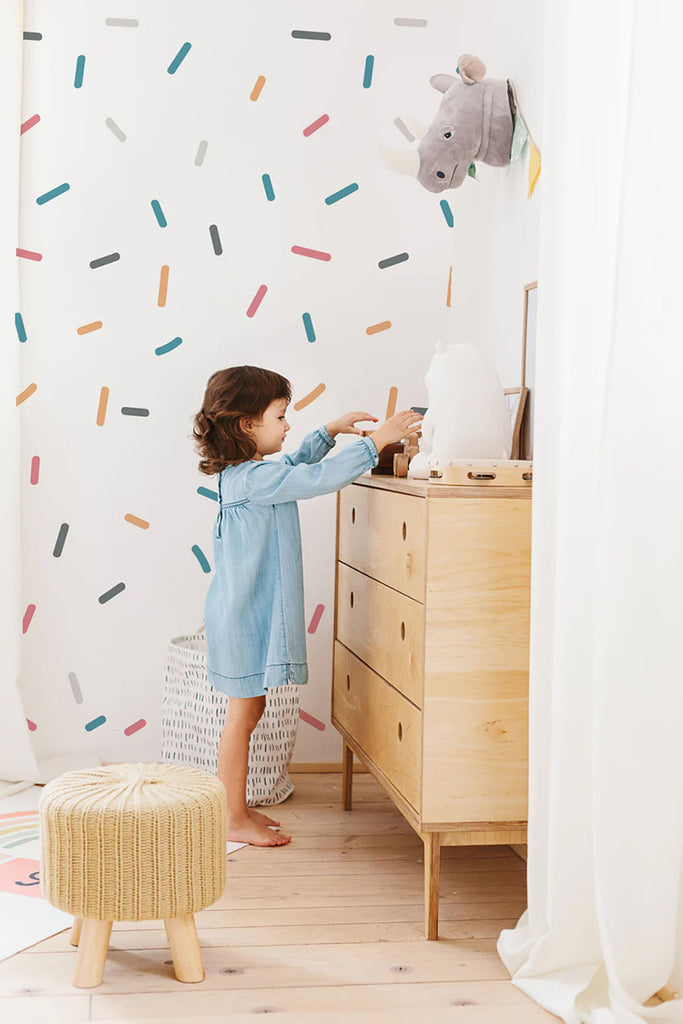 A lively child’s room with Confetti Shower, Pattern Wallpaper in Turquoise Maroon, creating a vibrant backdrop. A child in a blue dress interacts with a wooden dresser, while a whimsical stuffed animal head adorns the wall. A small stool adds to the room’s playful charm.