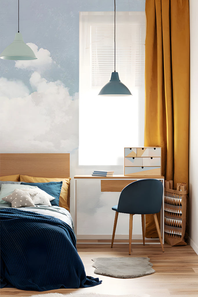 A tranquil bedroom adorned with Cuddle Clouds, Mural Wallpaper in Blue, depicting fluffy white clouds against a soothing blue sky. The room features a wooden bed with navy bedding, a modern desk and chair, mustard yellow curtains, and chic hanging lamps.
