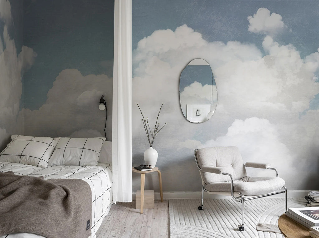 Serene bedroom featuring Cuddle Clouds, Mural Wallpaper in Blue. Fluffy white clouds on a soft blue sky create a calming atmosphere. Room includes a cozy bed, armchair, round mirror, and minimalist decor.