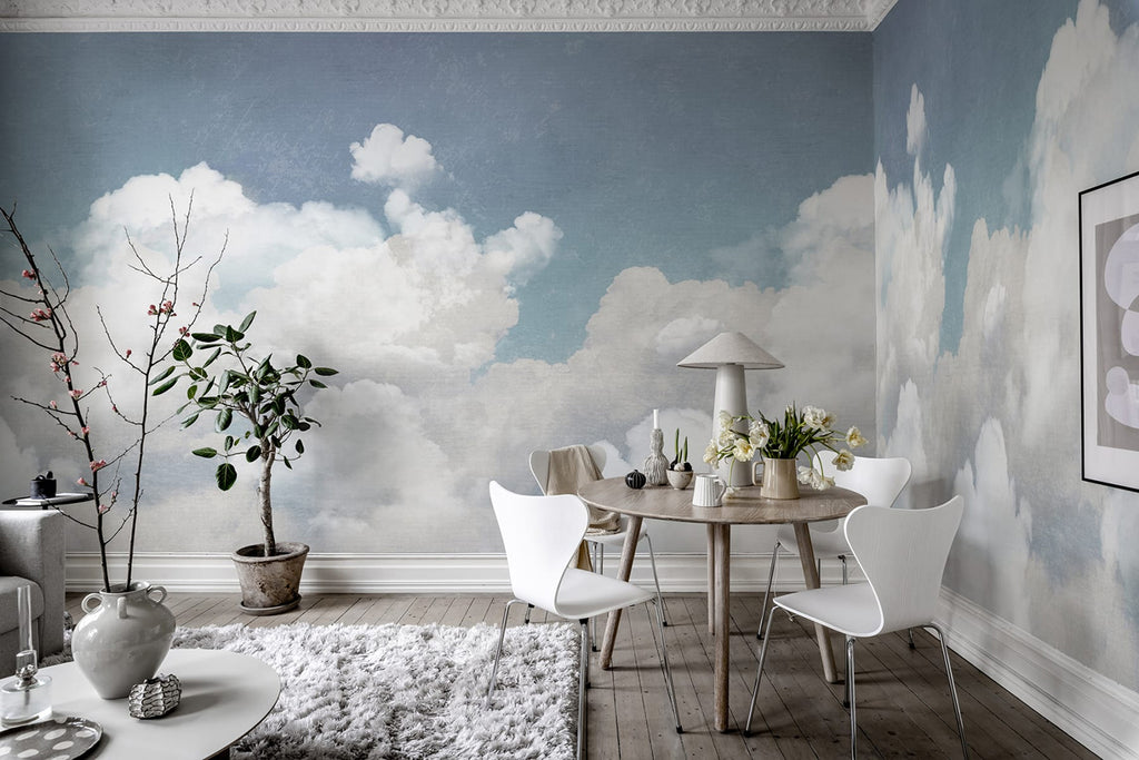 Serene room with Cuddle Clouds, Mural Wallpaper in Blue, featuring a soft blue sky with fluffy white clouds. The room includes a modern dining set, decorative plants, and chic lighting.