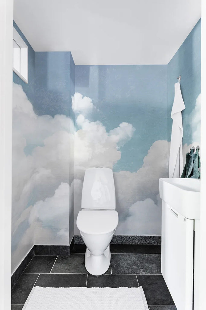 Serene bathroom with Cuddle Clouds, Mural Wallpaper in Blue, depicting a realistic sky and fluffy clouds. The peaceful atmosphere is enhanced by the toilet and sink against the calming backdrop.