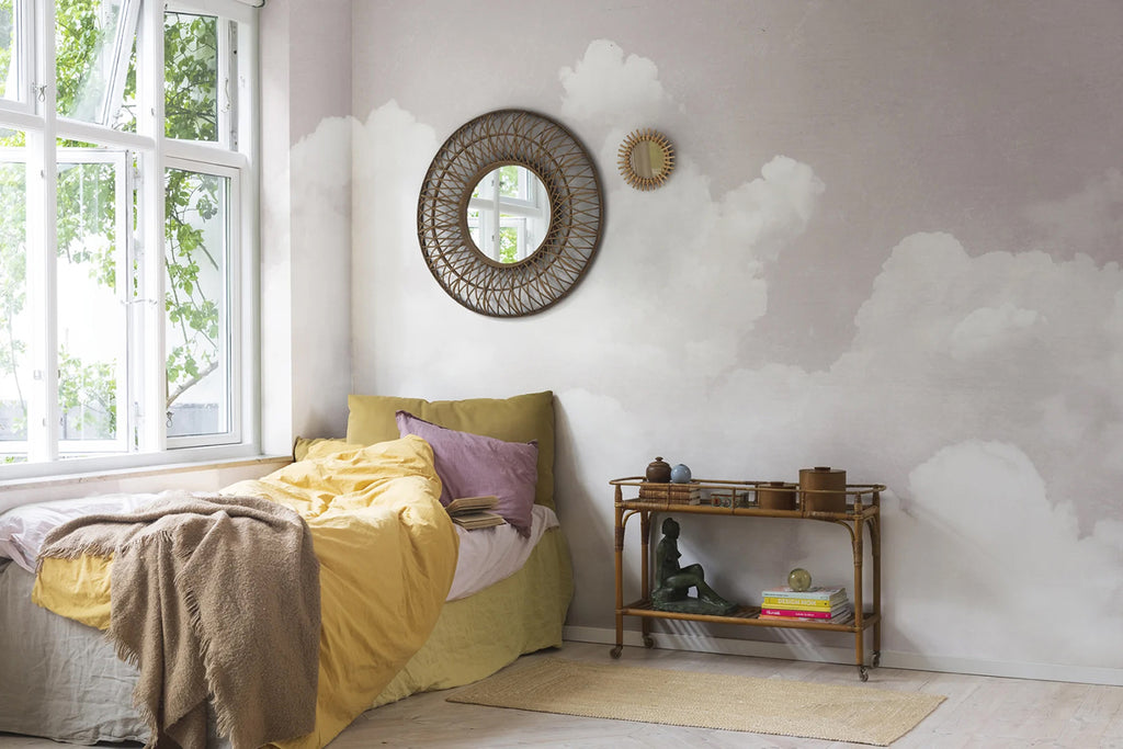 Cozy bedroom featuring Cuddle Clouds, Mural Wallpaper in Pink  with fluffy white clouds on a soft pink background. The room includes a bed with yellow and purple bedding, a round mirror, decorative items, and a window.