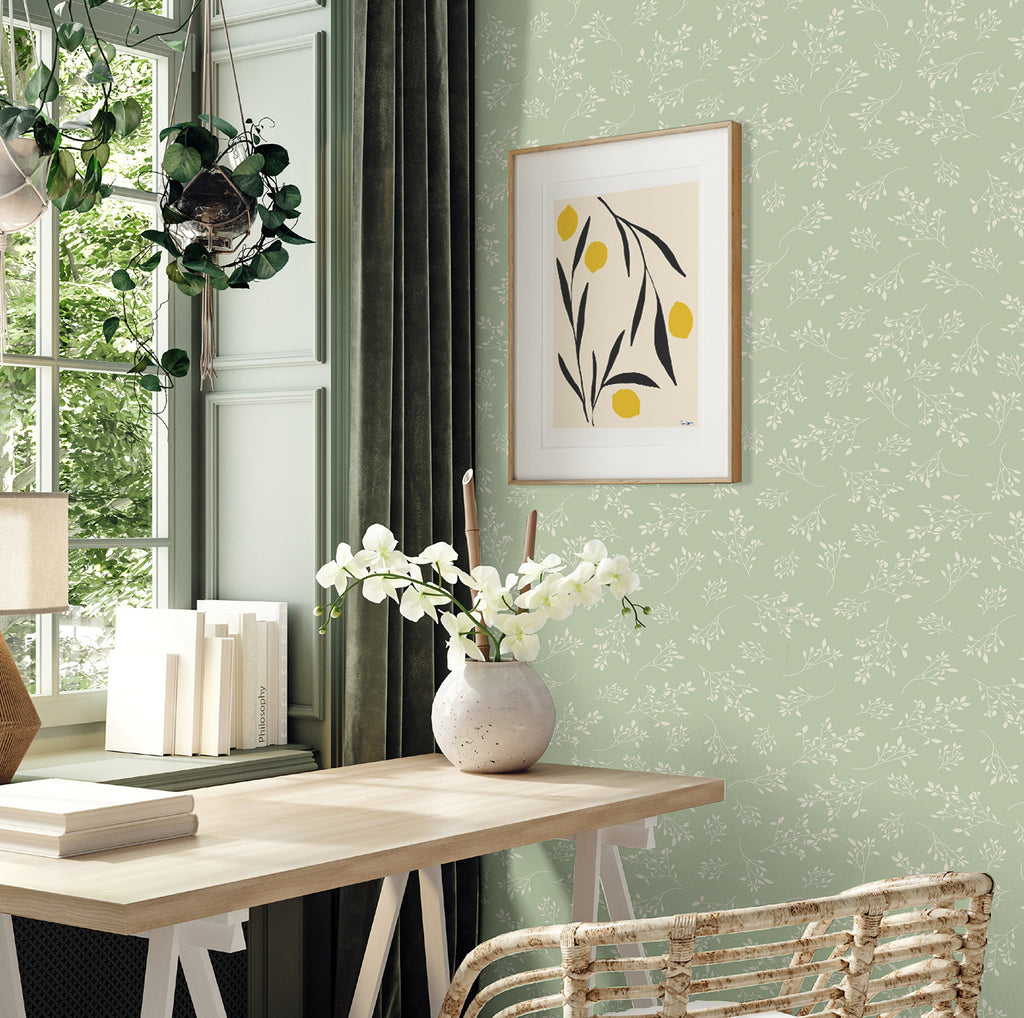 In an elegant interior, natural light filters through large windows, illuminating the subtle Delicate Foliages, Pattern Wallpaper in Green. The room boasts a wooden desk, a collection of books, and a rattan chair, creating an inviting atmosphere.