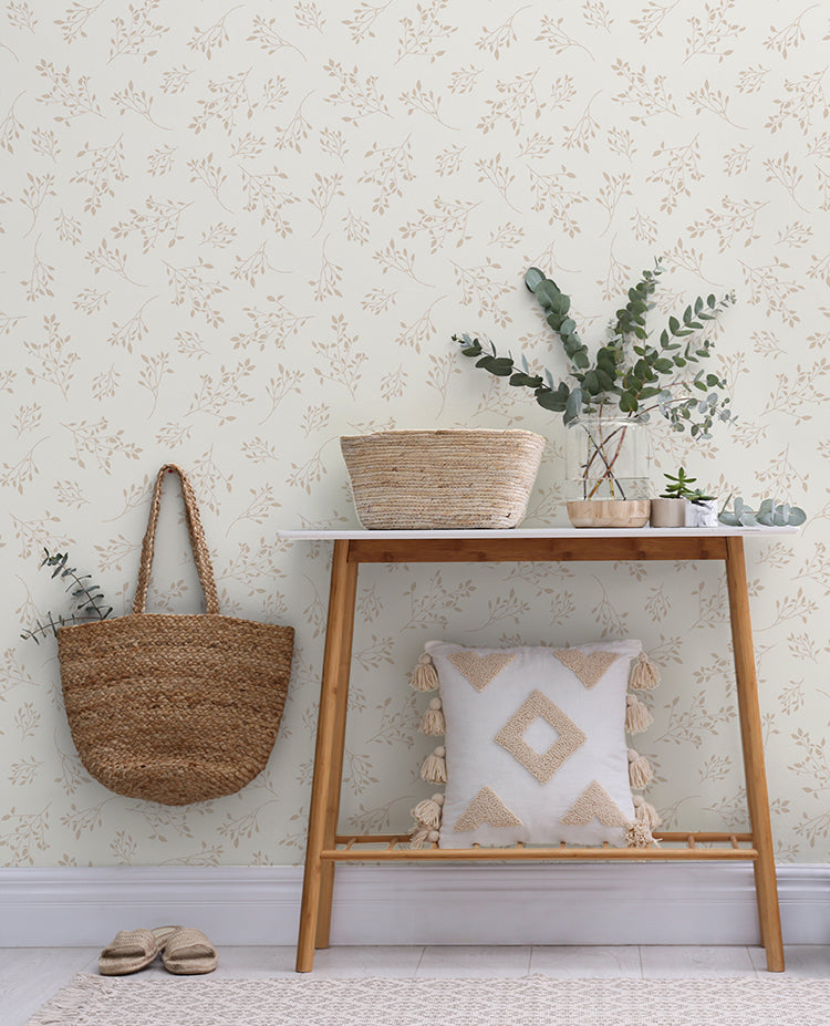 In a cozy corner, the Delicate Foliages, Pattern Wallpaper in Nude graces the walls with its subtle green and brown tones. The design evokes a sense of serenity, reminiscent of a tranquil forest. A wooden shelf adorned with a woven basket and greenery complements the wallpaper. Nearby, a cushioned bench invites relaxation, and a pair of slippers rest on the floor.