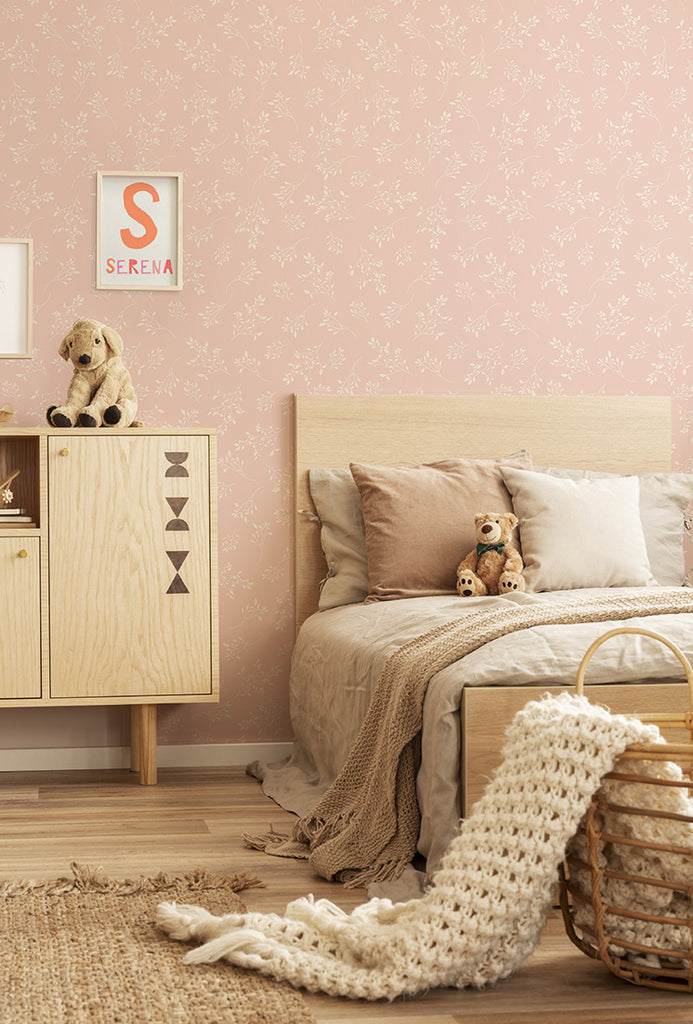 The serene bedroom features the Delicate Foliages, Pattern Wallpaper in Pink adorned with subtle botanical prints. The wooden bed, dressed in beige linens, pairs beautifully with a matching nightstand and a woven basket. Plush toys add a touch of coziness, while a framed ‘SERENA’ print on the wall personalizes the space.