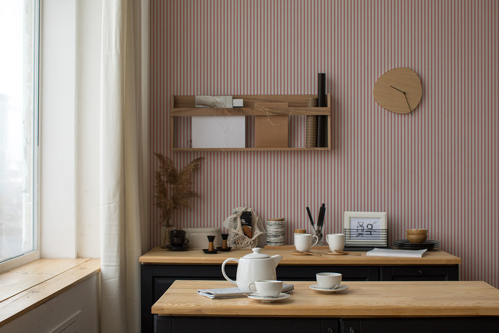 A serene workspace bathed in natural light from a window. A desk with neatly arranged art supplies sits under a wooden shelf holding books and decor, all against a wall with Delicate Stripes, Pattern Wallpaper in Blush Pink.