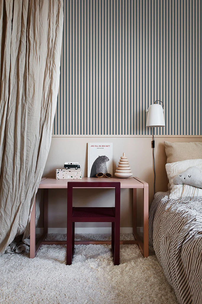 A welcoming bedroom bathed in soft light. A bed with striped linens sits next to a wooden bedside table with a lamp and book, all set against a wall featuring elicate Stripes, Pattern Wallpaper in Dark Blue.