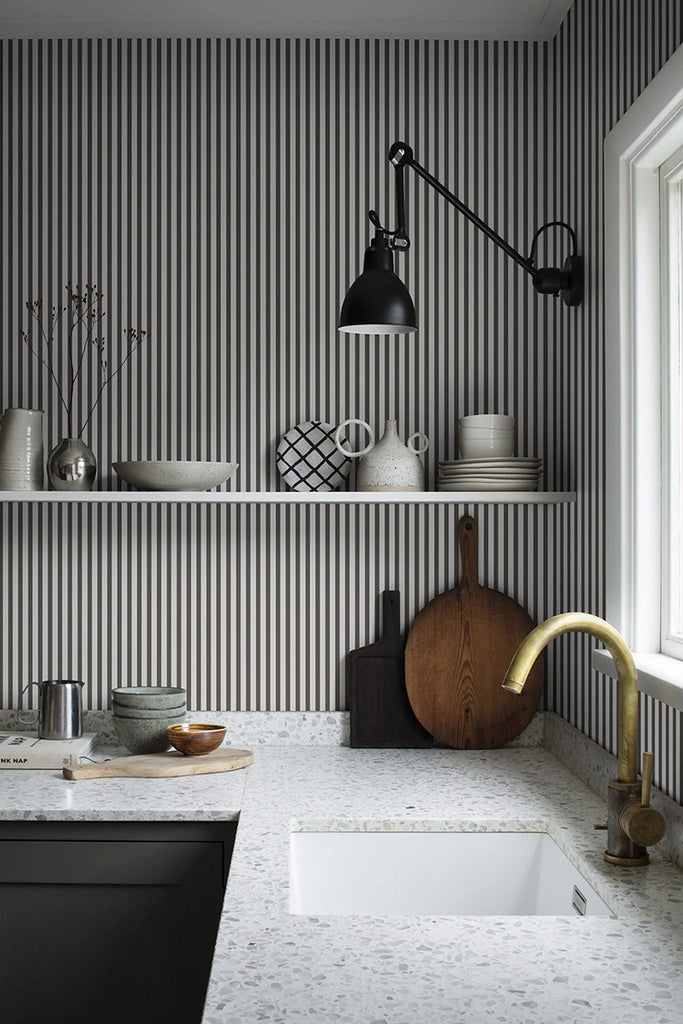 An elegant kitchen with a marble countertop and brass faucet, a black lamp hanging above white shelves displaying decor. All set against a wall featuring Delicate Stripes, Pattern Wallpaper in Dark Grey.