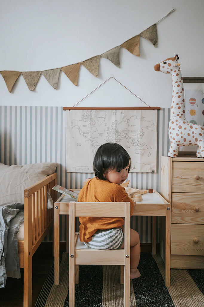 A kids room featuring a wooden dresser, bed and study table against the Delicate Stripes, Pattern Wallpaper in Grey Blue. A bunting drapes across the wall and a world map poster hangs on the wall.