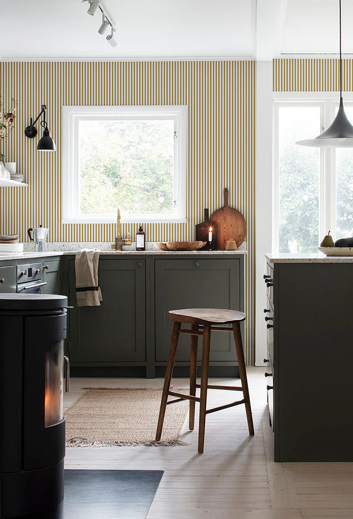 A modern kitchen bathed in natural light, featuring olive green cabinetry and a wooden countertop. A round stool sits near the counter, all set against a wall with Delicate Stripes, Pattern Wallpaper in Honey.