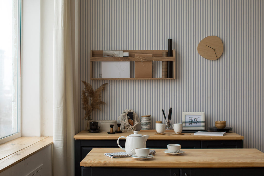 A serene workspace bathed in natural light from a window. A desk with neatly arranged art supplies sits under a wooden shelf holding books and decor, all against a wall with Delicate Stripes, Pattern Wallpaper in Light Blue.