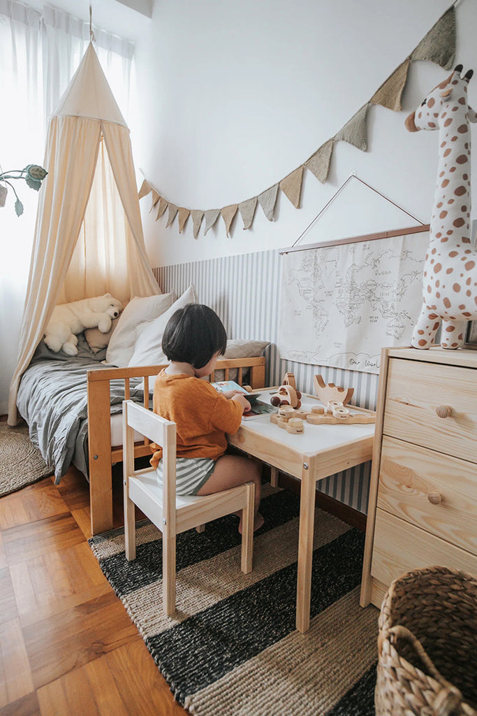 A child’s bedroom with a cozy ambiance, featuring a small wooden bed and desk. A child sits at the desk, engaged in an activity. Decorative elements like a canopy, bunting flags, and wall art add charm, all against a Delicate Stripes, Pattern Wallpaper in Light Grey.