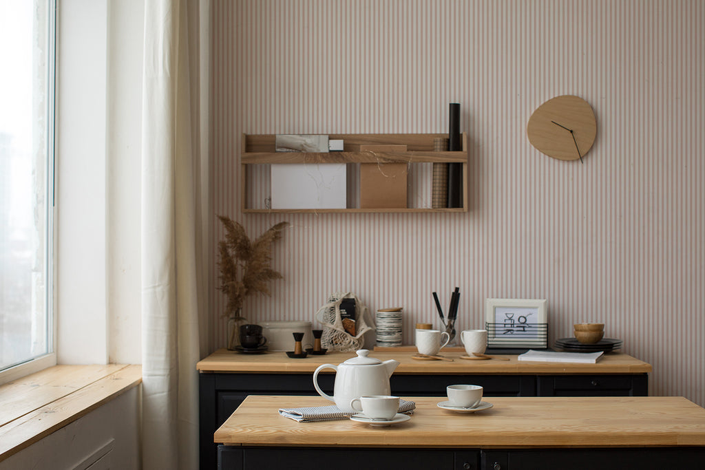 A serene workspace bathed in natural light from a window. A desk with neatly arranged art supplies sits under a wooden shelf holding books and decor, all against a wall with Delicate Stripes, Pattern Wallpaper in Nude.