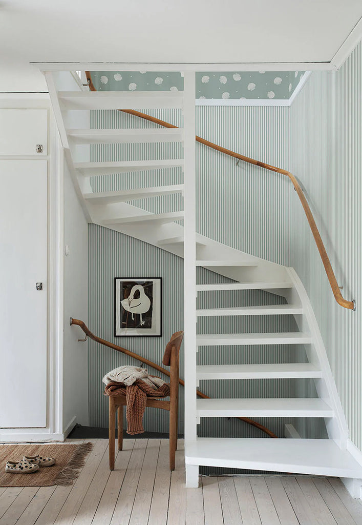 A serene room with a white staircase and wooden handrails, accented by a Delicate Stripes, Pattern Wallpaper in Sage. A framed artwork hangs on the wall, and a wooden chair with a draped blanket adds warmth.