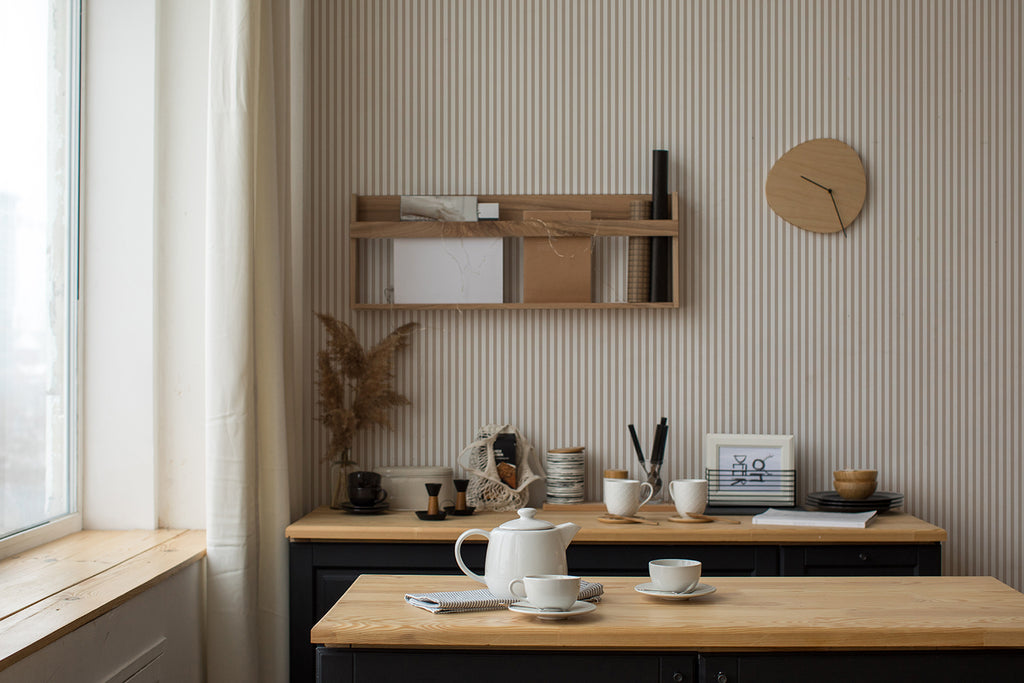 A serene workspace bathed in natural light from a window. A desk with neatly arranged art supplies sits under a wooden shelf holding books and decor, all against a wall with Delicate Stripes, Pattern Wallpaper in Sand.