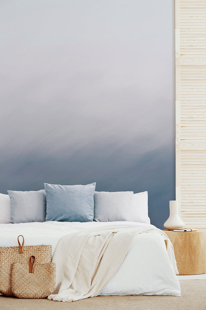 The Delilah Waves, Watercolor Wallpaper in Blue graces a serene bedroom. Its gradient from white to deep blue evokes calmness. The room features a bed adorned with white and light blue pillows, a cream blanket, and a woven basket beside it.