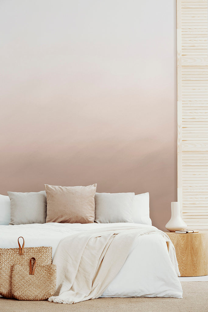 Delilah Waves, Watercolor Wallpaper in Sand graces a serene bedroom. Its gradient from white to soft brown evokes calmness. The room features a bed adorned with white and light brown pillows, a cream blanket, and a woven basket beside it.