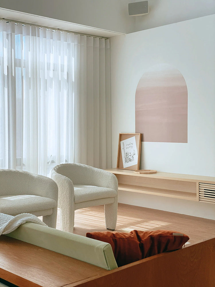 A sunlit room with sheer curtains, a cream armchair, and a wooden bench adorned with framed artwork. Dome Ombre Gradient, Wall Decals in Nude  adds a touch of brown above the bench.