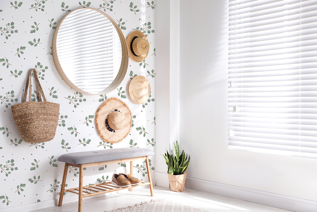 Serene interior with Eucalyptus Leaf, Wallpaper. Green leaves on white backdrop. Decor includes a round mirror, straw hats, woven bag, wooden bench with shoes, and a potted plant.